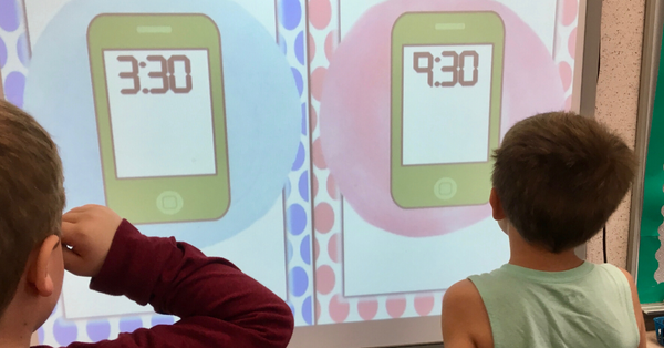 Two boys are facing a whiteboard. There is a slide projected on the whiteboard that has 2 green cell phones. One phone displays "3:30" in digital numbers and the phone on the right displays 9:30 in digital numbers. 