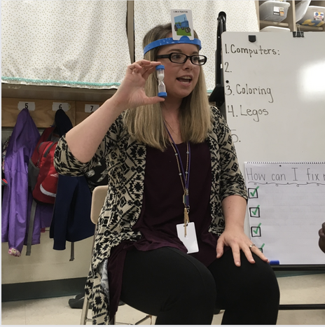 a photo of myself wearing a headbanz headband and card and holding up a sand timer for my students.