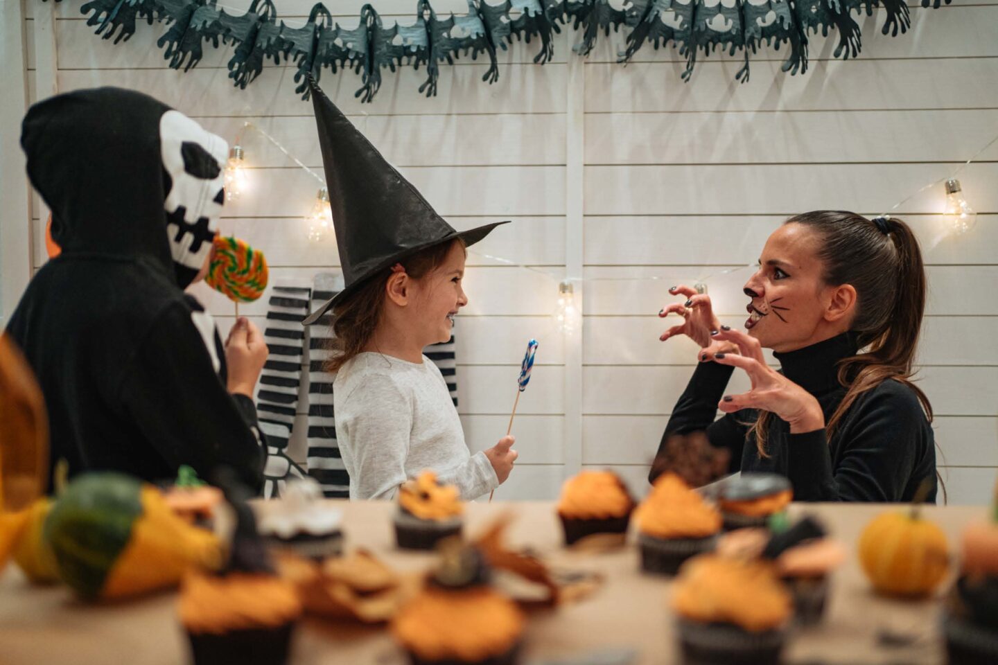A photo showing one of the Halloween classroom ideas of telling a group scary story. In the photo a boy in a mask and a girl in a witch hat are listening to a grownup woman dressed as a cat tell a scary story.